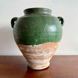 May include: A large, bulbous earthenware vessel featuring a wide mouth and two small side handles. The upper portion is coated in a glossy, deep green glaze, while the lower section remains exposed, showing a textured, light tan terracotta surface.