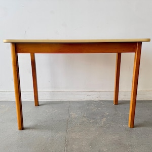 May include: A rectangular table featuring a light-coloured, smooth top surface with a thin edge, supported by four straight, medium-brown wooden legs. The frame connecting the legs is also made of the same medium-brown wood, creating a simple, functional structure with clean lines and a balanced, sturdy appearance.