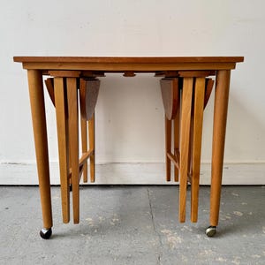 Op de afbeelding: Een vintage houten bijzettafel met een rechthoekig blad en opvouwbare bladeren. De tafel heeft vier afgeronde poten, waarvan twee met kleine wielen. De tafel is gemaakt van lichtbruin hout.