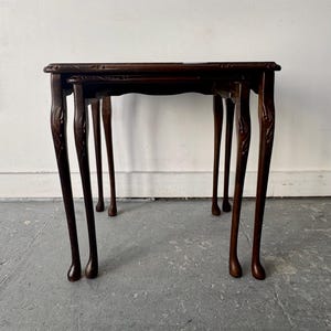 May include: Set of two antique dark brown nesting tables. The tables have carved details on the legs and edges. The legs are slender and curved, with a ball-and-claw foot design. The tables are of different sizes and can be stored together.
