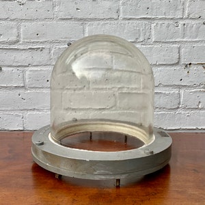 Könnte beinhalten: Eine klare Glaskuppel mit grauem Metallsockel. Die Kuppel ist abgerundet und sitzt auf einem kreisförmigen Sockel mit kleinen Füßen. Der Sockel hat eine hellgraue Oberfläche. Der Hintergrund ist eine weiße Backsteinmauer.