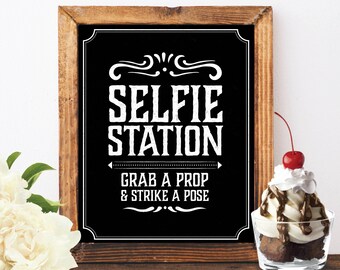 May include: A black chalkboard sign with white text that reads "Selfie Station Grab a Prop & Strike a Pose". The sign is framed in wood and is sitting on a wooden surface.