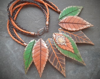 May include: A necklace featuring a braided leather cord and five leaf-shaped pendants. The leaves are crafted from polymer clay and are painted in shades of green, brown, and orange. The central leaf has a decorative metal embellishment with a light-colored stone.