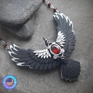 Pode incluir: Um colar com pingente de corvo preto com uma grande pedra preciosa vermelha oval no centro. As asas do corvo estão estendidas, com detalhes prateados nas penas. A corrente do colar apresenta pequenas contas vermelhas.