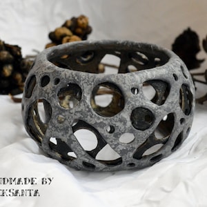 Könnte beinhalten: Ein grauer Keramik-Pflanztopf mit einem einzigartigen, durchbrochenen Design. Der Topf hat eine runde Form und ist aus einem strukturierten Material gefertigt. Er ist handgefertigt von Aleksanta.