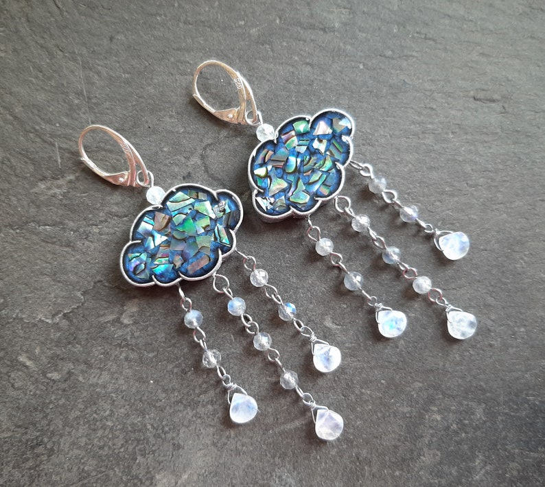 May include: A pair of silver earrings featuring a blue and green iridescent cloud-shaped charm with a dangling chain of white moonstone beads.