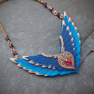 May include: A blue and gold winged necklace with a red gemstone heart pendant. The necklace is made of polymer clay and features a detailed design.