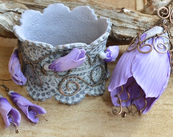 Puede incluir: Un conjunto de joyas en forma de flor de color lavanda, que incluye un brazalete, pendientes y un colgante. Las joyas están hechas de arcilla polimérica y presentan delicados detalles de alambre.