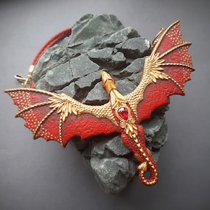 May include: A red and gold dragon pendant with a red gemstone, resting on a gray rock. The dragon has detailed wings and a red cord necklace. The pendant is a fantasy-themed accessory.