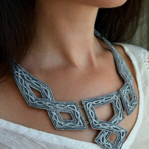 Puede incluir: Un collar de fieltro gris con un dise&ntilde;o geom&eacute;trico. El collar est&aacute; hecho de m&uacute;ltiples hebras de fieltro que se tejen juntas para crear un patr&oacute;n &uacute;nico e intrincado. El collar es una pieza llamativa que a&ntilde;adir&iacute;a un toque de elegancia a cualquier atuendo.