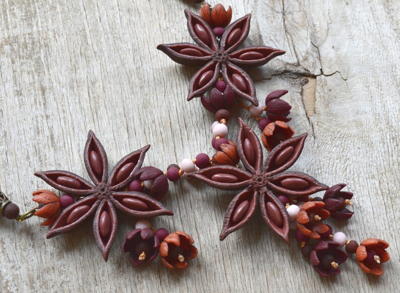 May include: A brown star anise necklace with burgundy and orange flower accents. The necklace is made of polymer clay and is strung on a brown cord.