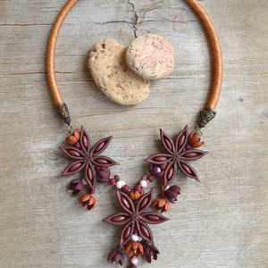 May include: A brown leather cord necklace with a star anise pendant. The pendant is made of brown and burgundy polymer clay and features a cluster of star anise flowers. The necklace is accented with small brown and white beads.
