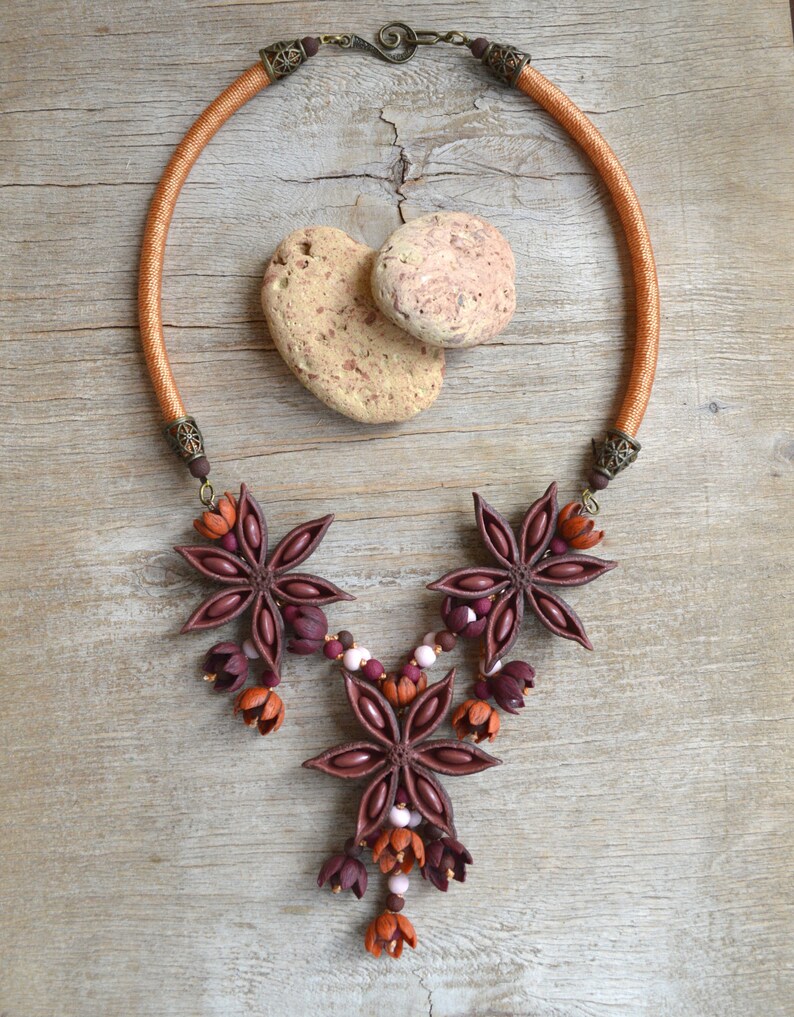 May include: A brown leather cord necklace with a star anise pendant. The pendant is made of brown and burgundy polymer clay and features a cluster of star anise flowers. The necklace is accented with small brown and white beads.