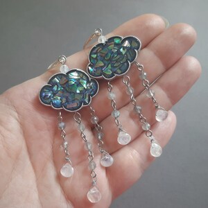 May include: A pair of silver cloud-shaped earrings with iridescent blue and green abalone shell inlays. Each earring has a silver chain with multiple clear teardrop-shaped crystals.