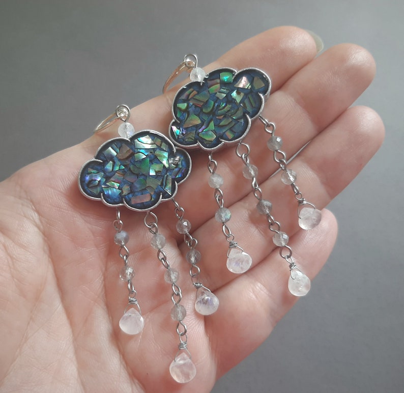 May include: A pair of silver cloud-shaped earrings with iridescent blue and green abalone shell inlays. Each earring has a silver chain with multiple clear teardrop-shaped crystals.