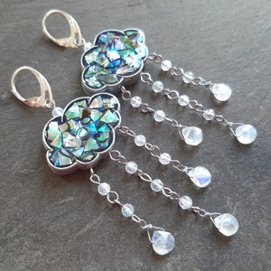 May include: A pair of silver earrings with a cloud-shaped design. The clouds are made of iridescent abalone shell and are accented with dangling crystal beads.