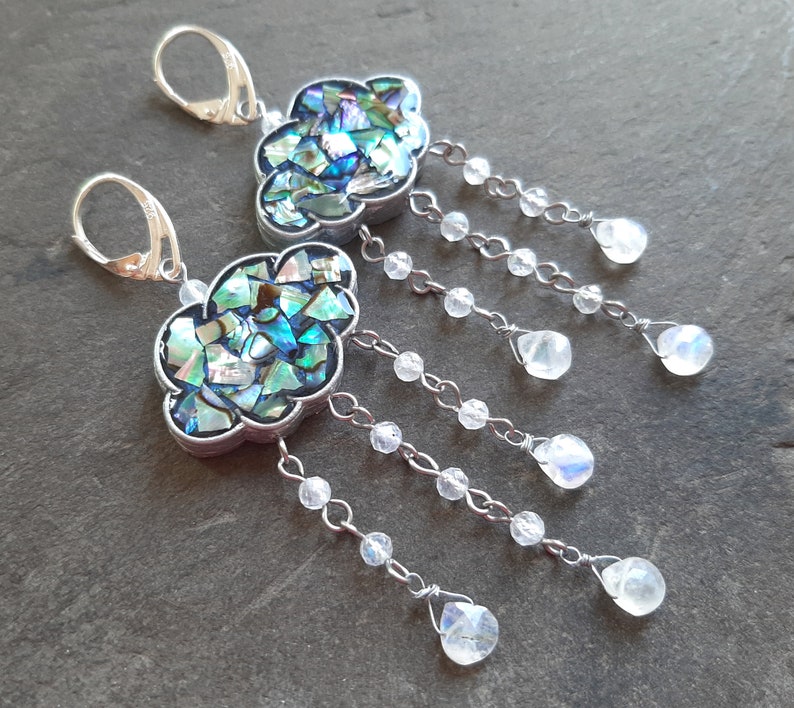 May include: A pair of silver earrings with a cloud-shaped design. The clouds are made of iridescent abalone shell and are accented with dangling crystal beads.