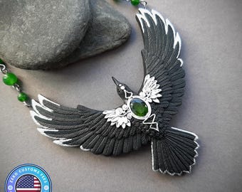 May include: A black raven pendant necklace with silver-colored accents and a green gemstone. The raven's wings are outstretched, and the necklace features green beads. The pendant is set against a gray background.