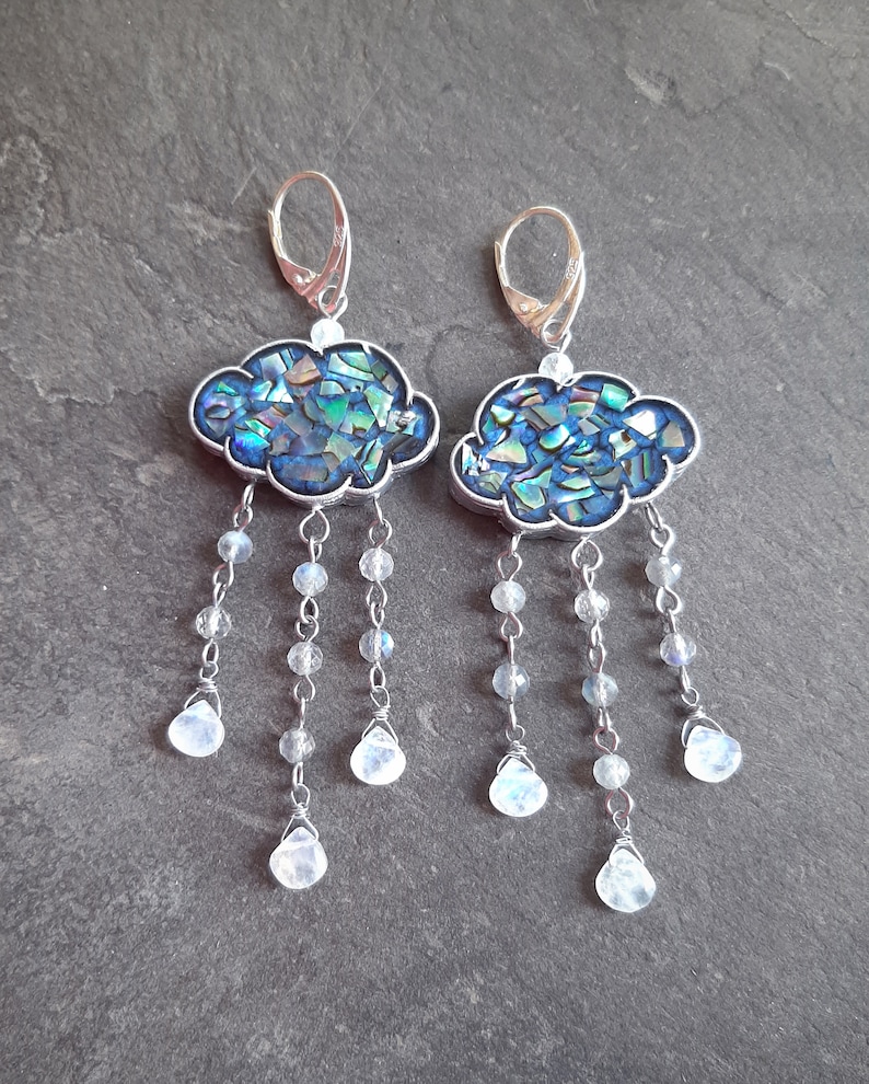 May include: A pair of silver earrings with a cloud-shaped charm made of iridescent blue and green abalone shell. Each cloud charm has a silver chain with five dangling moonstone beads.