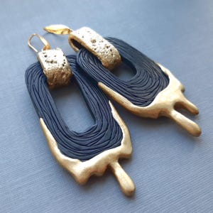 以下が含まれることがあります： 幾何学模様のブラックとゴールドのステートメントイヤリング。イヤリングはポリマークレイ製で、テクスチャ加工が施されています。