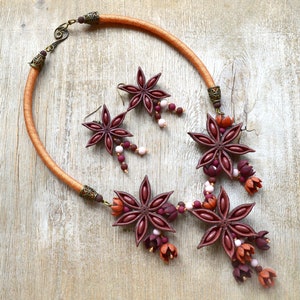 May include: A brown leather cord necklace with three star anise charms and pink and burgundy beads. The necklace has a brown leather cord and a silver clasp. The star anise charms are made of polymer clay and are painted in shades of brown, burgundy, and pink.