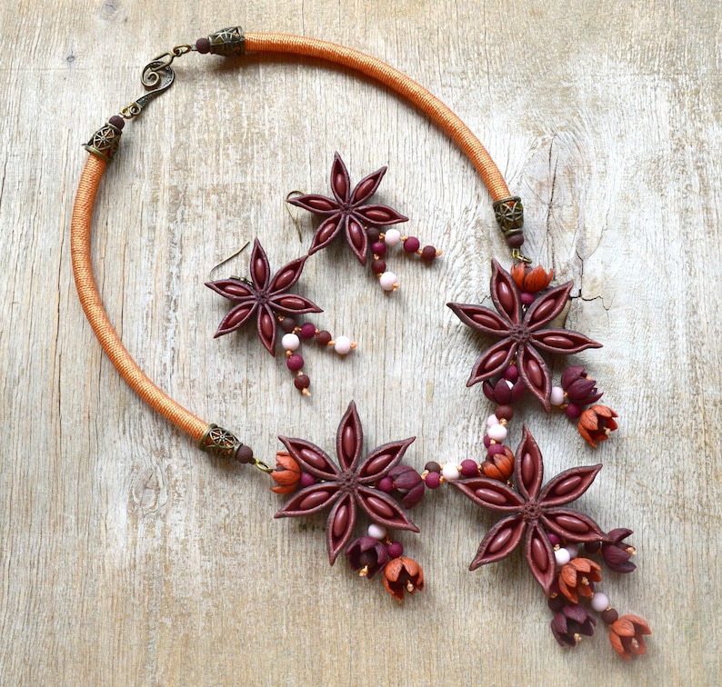 May include: A brown leather cord necklace with three star anise charms and pink and burgundy beads. The necklace has a brown leather cord and a silver clasp. The star anise charms are made of polymer clay and are painted in shades of brown, burgundy, and pink.
