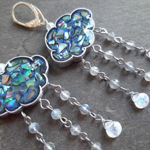 May include: A pair of silver earrings with a cloud-shaped pendant made of iridescent blue and green mosaic tiles. Each cloud is attached to a silver chain with multiple dangling moonstone beads.