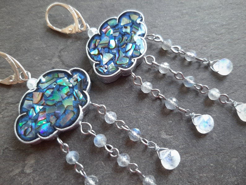 May include: A pair of silver earrings with a cloud-shaped pendant made of iridescent blue and green mosaic tiles. Each cloud is attached to a silver chain with multiple dangling moonstone beads.