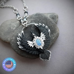 May include: A silver necklace featuring a black raven pendant with outstretched wings. The raven has a blue gemstone center and silver accents. The necklace chain includes black beads and a circular pendant holder.