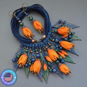 Könnte beinhalten: Ein handgefertigtes Halsketten- und Ohrring-Set mit orangefarbenen Blumenanhängern, blauen Perlen und grünen Blattakzenten. Die Halskette besteht aus mehreren Strängen blauer Kordel. Die Ohrringe haben passende orangefarbene Blumenanhänger und grüne Perlen. Ein einzigartiges Set.