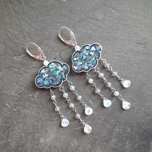 May include: A pair of silver earrings featuring a blue and green iridescent cloud design with a dangling chain of white moonstone beads.