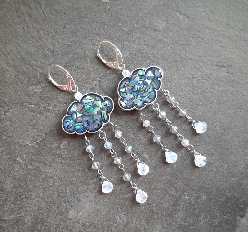 May include: A pair of silver earrings featuring a blue and green iridescent cloud design with a dangling chain of white moonstone beads.