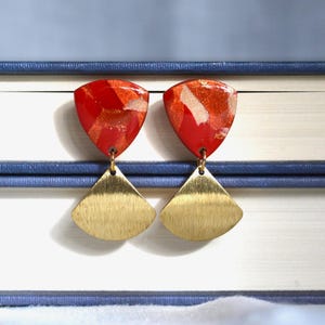 Può includere: Orecchini con parti superiori triangolari rosse e dorate e parti inferiori dorate a forma di ventaglio. Sono presentati su uno sfondo bianco, con un libro blu sullo sfondo.