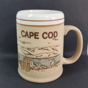 May include: A beige ceramic mug with a handle, featuring the words "CAPE COD" in brown lettering. The mug has a coastal scene with seagulls and a shoreline design. The rim is white with a brown accent.