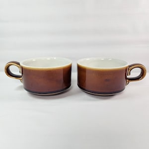 May include: Two brown ceramic coffee cups with a rounded handle. The cups have a glossy finish and a white interior. The cups are sitting on a white surface.