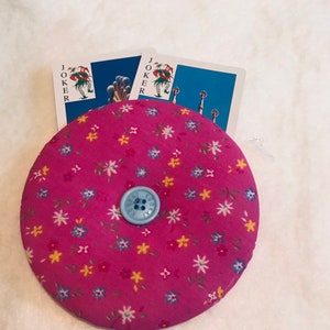 May include: A circular, pink pin cushion with a floral design and a light blue button in the middle. Two playing cards with "JOKER" printed on them are behind the pin cushion. The pin cushion is approximately 10 cm in diameter.