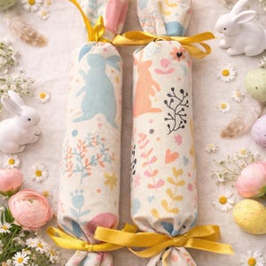 May include: Two fabric Easter crackers with bunny and floral patterns, tied with yellow ribbons. The crackers are surrounded by Easter eggs, white ceramic bunnies, and flowers, creating a festive spring scene.