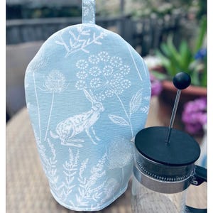 May include: A pale blue tea cosy featuring a white rabbit and floral design, alongside a glass French press coffee maker. The cosy has a hanging loop. The French press has a black lid and handle.