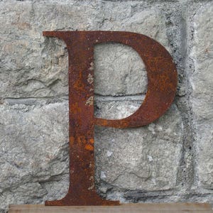 May include: A rusty metal letter P, standing upright against a grey stone wall. The letter is large and has a simple, classic font.
