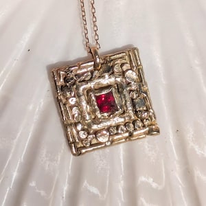 May include: A gold-coloured pendant necklace with a square, textured design. The pendant features a central, square-cut, red gemstone. The necklace chain is delicate and matches the pendant's colour. The jewellery is displayed on a white surface.