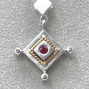 May include: Silver pendant with a diamond-shaped design. The pendant features a central square with a ruby gemstone, framed by gold rope detailing. Small, round, clear gemstones are set at each corner of the diamond shape.