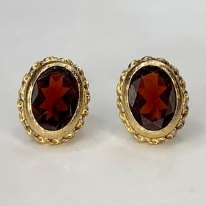 May include: A pair of gold-toned stud earrings. Each earring features an oval, faceted, dark red gemstone set within a decorative, twisted gold frame. The gemstones have a star-like pattern in the center.