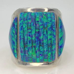 Puede incluir: Un anillo de plata con una piedra central de ópalo rectangular, iridiscente azul y verde. El ópalo está segmentado con líneas verticales, y el anillo tiene detalles de ópalo redondeados a cada lado.