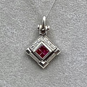 May include: A silver pendant necklace featuring a square, ruby-colored gemstone at the center. The pendant is surrounded by small, clear gemstones and a geometric design. The necklace is displayed against a gray background.