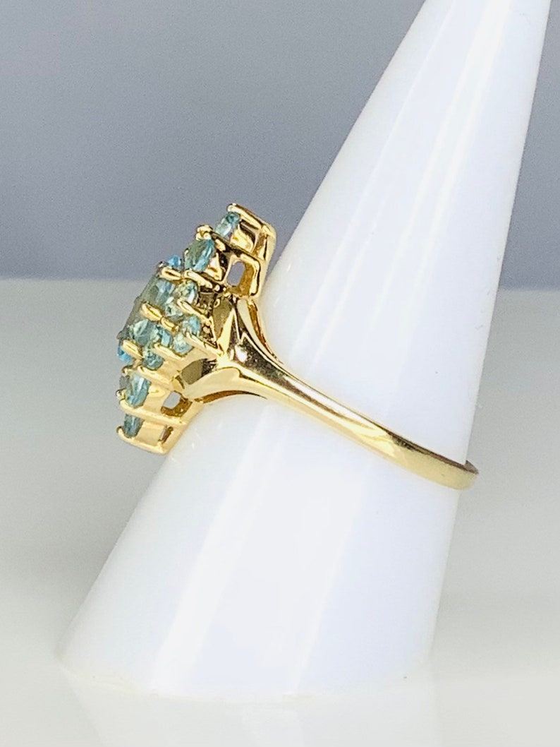 May include: A gold ring with a cluster of light blue gemstones. The ring is set on a white display stand.