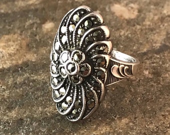 May include: A silver-toned ring with a large, oval-shaped face. The face features a central flower design surrounded by a fan-like pattern, all embellished with small, dark stones. The ring's band has decorative etched details.