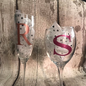 May include: Two clear glass wine glasses with a white polka dot pattern. The glass on the left has a copper-coloured letter "R" on it. The glass on the right has a pink letter "S" on it.
