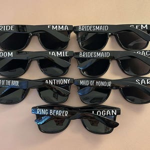 May include: Set of ten black sunglasses with white text on the frames. The text includes "Bride", "Groom", "Maid of Honour", "Bridesmaid", "Brother of the Bride", "Ring Bearer", "FMMA", "JAMIE", "ANTHONY", "LOGAN", "SEM", "PACE", and "SARA".