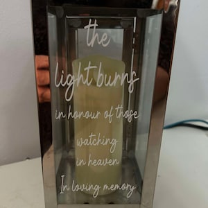 May include: A silver metal lantern with a glass panel featuring the text "the light burns in honour of those watching in heaven in loving memory".