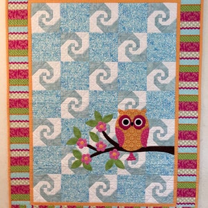 May include: A quilt with a blue and white swirl pattern and a pink owl sitting on a branch with pink flowers. The quilt has a pink, green, and orange border.